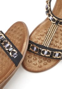 Zwarte slipper-sandalen met gouden kettingversiering, gestructureerde gouden binnenzool, platte zool en decoratieve zwart-grijze accenten.
