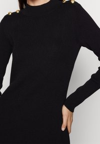 Černé splývavé pletené šaty s vysokým výstřihem, zdobené zlatými knoflíky na rameni. Dlouhé rukávy a přiléhavý tvar umocňují design.