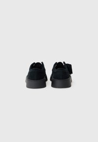 Sorte semskede sneakers med lav profil, rund tå og teksturert overdel. Har sorte gummisåler og en løftestrop bak.
