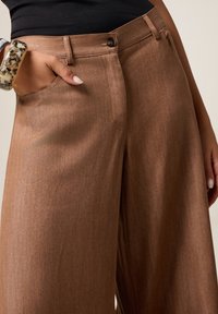 Pantalons évasés marron en tissu texturé avec une fermeture à bouton et des poches latérales. Dotés d'une finition lisse et de lignes épurées.
