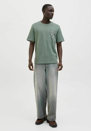 Grünes T-Shirt mit kurzen Ärmeln und einer gemusterten Tasche, kombiniert mit lockeren, hellblauen Jeans und dunklen Stiefeln. Einfaches, lässiges Design.
