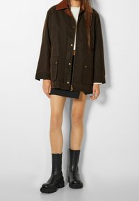 Brun canvasjakke med brun fløjlskrave, der har guld trykknapper, to forlommer og en let oversized pasform. Sorte ankelstøvler.