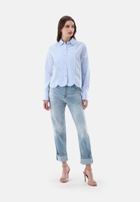 Camicia azzurra con colletto e orlo scalloppato, abbinata a jeans azzurri con risvolti e sandali con tacco nudo su uno sfondo semplice.