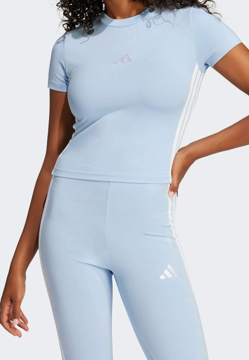 Ljusblå träningsöverdel och matchande leggings i ett mjukt tyg; har tre vita ränder på sidorna och en liten logotyp på bröstet.