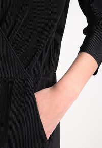 Svart plisserad klänning med V-ringning. Har långa ärmar och en sidficka. Materialet verkar vara texturerat med vertikala linjer.