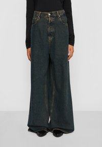 Persoon in hoge taille, wijde donkerblauwe jeans met zichtbare stiksels, zwart long-sleeve top en zwarte schoenen tegen een effen achtergrond.