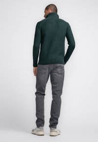 Donkergroene chunky gebreide coltrui met een geribbelde onderrand, gecombineerd met grijze spijkerbroek en lichtgekleurde sneakers.