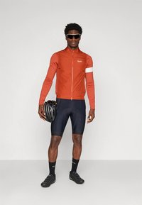 Mannelijke fietser in een oranje longsleeve shirt en zwarte fietsshorts, met een zwarte helm in zijn hand, en gekleed in zwarte sokken en fietsschoenen.