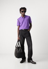 Μωβ πόλο μπλουζάκι με μαύρες λεπτομέρειες, μαύρα τζιν, μαύρα παπούτσια και μαύρη τσάντα. Μοντέλο που φοράει γυαλιά ηλίου, στέκεται μπροστά από ουδέτερο φόντο.