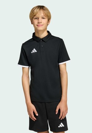 Ragazzo adolescente biondo in piedi, indossa una polo nera Adidas e pantaloncini con loghi bianchi, su uno sfondo chiaro uniforme.