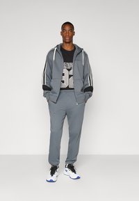 Šedý športový outfit, ktorý sa skladá z mikiny na zips s bielymi pruhmi a zodpovedajúcich teplákov. Čierne tričko s výrazným šedým číselným potlačou. Biele tenisky.