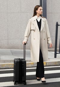 Trench coat beige de doble botonadura con solapas anchas, pantalones negros y una maleta negra con ruedas, en un paso de peatones. Textura suave, corte ajustado.