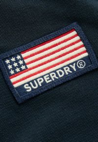 Námořnická látka s vyšitým logem, které zobrazuje stylizovanou americkou vlajku a slovo "SUPERDRY" v bílé barvě, zdůrazňující červené a bílé pruhy.