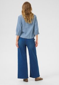 Chemise en denim bleu clair à manches 3/4, poignets à un bouton ; associée à un jean bleu foncé à jambe large et à des chaussures plates marron.