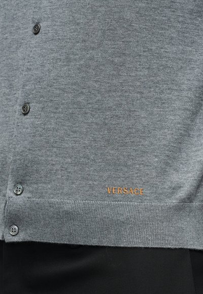 Gros plan sur un cardigan gris boutonné avec un ourlet côtelé et un petit logo doré "VERSACE" brodé près du bord inférieur.