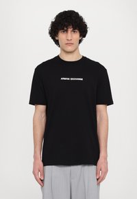 Svart bomull T-skjorte med "ARMANI EXCHANGE" logo i hvitt, kortermet, rund hals og glatt tekstur. Brukt sammen med grå plisserte bukser.