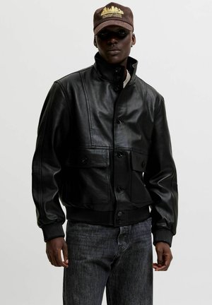 Muž nosi čiernu koženú bundu s gombíkmi vpredu a s klopovými vreckami, v kombinácii s tmavými džínami a hnedou šiltovkou so zlatou výšivkou.