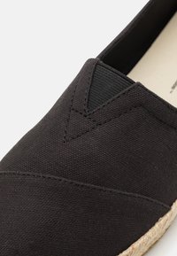 Botte slip-on en toile noire avec une finition texturée, présentant des accents triangulaires et une semelle en jute tressé naturel pour plus de détails.