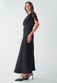 Robe maxi noire avec des manches courtes, corsage ajusté et jupe fluide. Présente une texture lisse et une fente sur le côté, assortie à des chaussures à talons pointus.