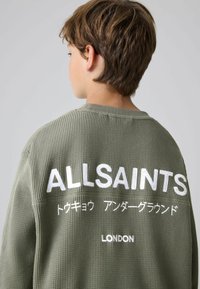 Felpa verde con texture a waffle, caratterizzata da una grande scritta bianca ricamata sul retro, che presenta "ALLSAINTS," testo giapponese e "LONDON."