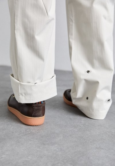 Weiße, strukturierte Hosen mit aufgerollten Säumen; braune Sneakers mit Wildleder- und Gummielementen, ausgestattet mit einem mehrfarbigen Streifen an der Ferse.