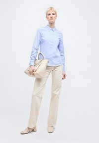 Γυναίκα με ανοιχτό γαλάζιο πουκάμισο με κουμπιά και μπεζ παντελόνι, κρατώντας μια μπεζ τσάντα, στέκεται μπροστά σε ένα καθαρό λευκό φόντο.