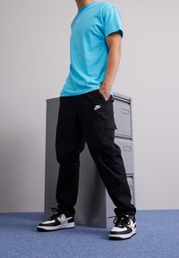Hombre que lleva pantalones cargo negros de Nike, zapatillas negras y blancas de Nike y una camiseta azul brillante, de pie cerca de un archivo gris.