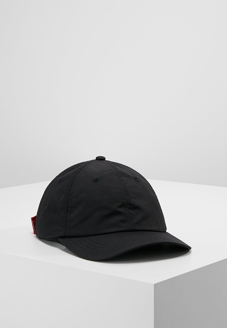 Casquette de baseball noire avec une visière incurvée et des panneaux cousus, posée sur un piédestal blanc sur fond gris.
