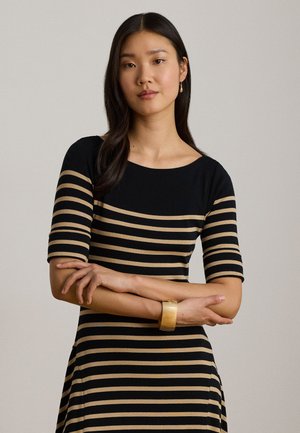 Femme aux longs cheveux foncés portant une robe rayée noire et beige, un bracelet en or et une boucle d’oreille pendante, debout les bras croisés devant un fond neutre.