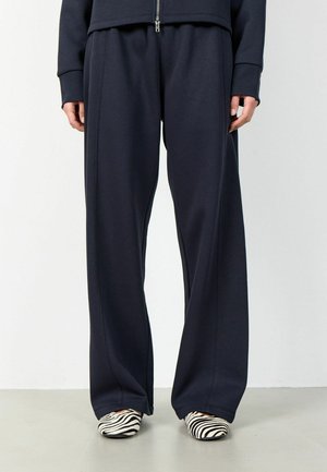 Oseba, ki nosi ohlapne črne hlače in črno-belo čevlje z zebra vzorcem, stoji na svetlo sivi tla ob beli steni.