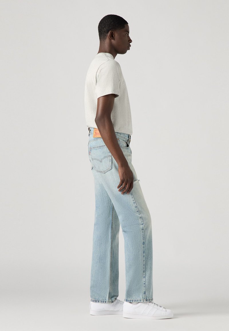 T-shirt gris clair, jeans droit bleu avec un délavage, et baskets blanches. Les jeans présentent une étiquette en cuir au dos.
