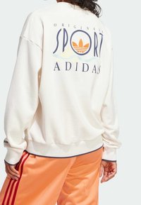 Cream sweatshirt with a large graphic reading "SPORT" and "ADIDAS" on the back, paired with orange track pants featuring red stripes.