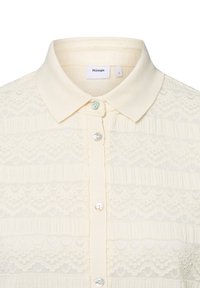 Cremfarbene Bluse mit Kragen, ausgestattet mit strukturierten Spitzenmustern und drei Perlenknöpfen auf der Vorderseite zum Schließen. Markenetikett sichtbar.