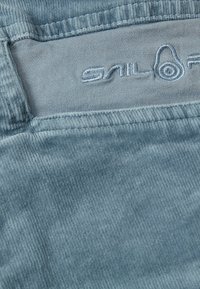 Vaaleansininen corduroy-kangas, jossa on pehmeä texture ja brodeerattu logotagi. Kankaassa on pystysuoraa röpelöä ja hieno kiilto.