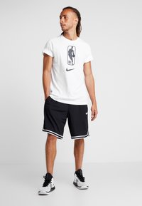 Camiseta blanca con gráfico de la NBA y logo de Nike, combinada con pantalones cortos de baloncesto negros con rayas blancas y logo de Nike. Zapatillas deportivas negras.