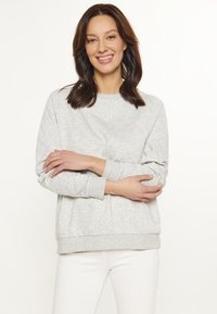Felpa grigia chiara in tessuto morbido e strutturato con scollo rotondo, polsini e orlo a costine. Indossata con pantaloni bianchi, braccia incrociate.