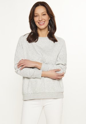 Lys grå sweatshirt i blødt, struktureret stof med rund halsudskæring, ribbede manchetter og kant. Båret med hvide bukser, armene korslagte.