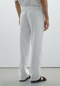 Pantalons en lin blancs à coupe décontractée, avec une taille élastique et une poche arrière, associés à des sandales noires ouvertes.