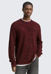Jonge man in een gestructureerde donkerrode trui over een wit shirt met grijze spijkerbroek, hand in zijn zak, staande tegen een effen lichte achtergrond.
