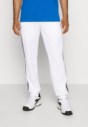 Uomo con pantaloni da ginnastica bianchi con strisce nere laterali, scarpe da ginnastica bianche e nere e maglietta blu, in piedi su uno sfondo semplice.