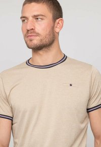 T-shirt beige à manches courtes avec un col rond rayé bleu marine et rouge, comportant une petite poche sur la poitrine avec un détail de bouton. Texture de tissu douce.