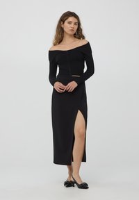 Top nero a spalla scoperta in maglia a costine con zip frontale, abbinato a una gonna maxi nera con uno spacco laterale. La modella indossa scarpe flat nere.