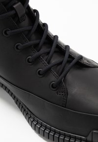 Zapatilla de cuero negra con suela de goma gruesa y cordones negros redondos pasados por ojales de metal sobre fondo blanco.