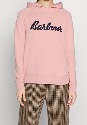 Person trägt pinkfarbenen Kapuzenpullover mit "Barbour"-Schriftzug und braune Hosen mit Hundeknochenmuster, steht vor einfarbigem Hintergrund.