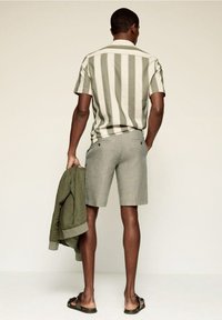 Uomo che indossa una camicia a maniche corte a righe verticali verdi e bianche, pantaloni corti grigi, sandali neri, con una giacca verde oliva in mano, in piedi davanti a uno sfondo neutro.