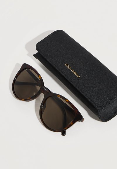 Γυαλιά ηλίου με σχέδιο χελώνας και σκουρόχρωμους φακούς δίπλα σε ένα μαύρο ορθογώνιο σκληρό θήκη με την επωνυμία Dolce & Gabbana πάνω σε λευκή επιφάνεια.