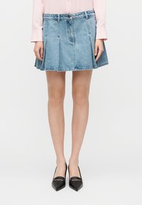 Μίνι φούστα από τζιν με πιέτες, ανοιχτό μπλε ξέβαμμα, ζωνάρι στη μέση με κουμπί, με ωμό τελείωμα, συνδυασμένη με μαύρες ψηλοτάκουνες μυτερές γόβες.