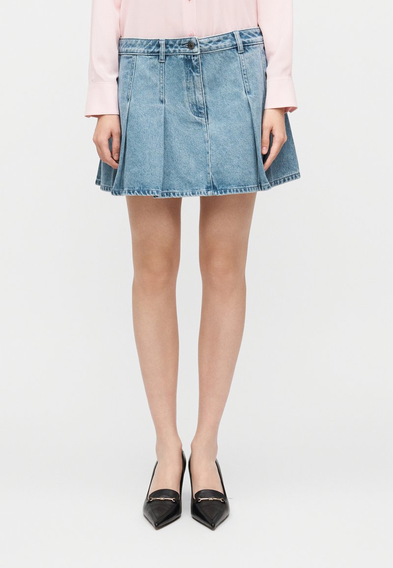 Μίνι φούστα από τζιν με πιέτες, ανοιχτό μπλε ξέβαμμα, ζωνάρι στη μέση με κουμπί, με ωμό τελείωμα, συνδυασμένη με μαύρες ψηλοτάκουνες μυτερές γόβες.