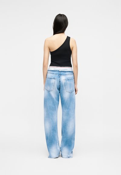 Γυναίκα με μακριά σκούρα μαλλιά που φοράει μαύρο τοπ με έναν ώμο και άνετο ξεθωριασμένο ανοιχτό μπλε τζιν, στέκεται με την πλάτη σε απλό φόντο.