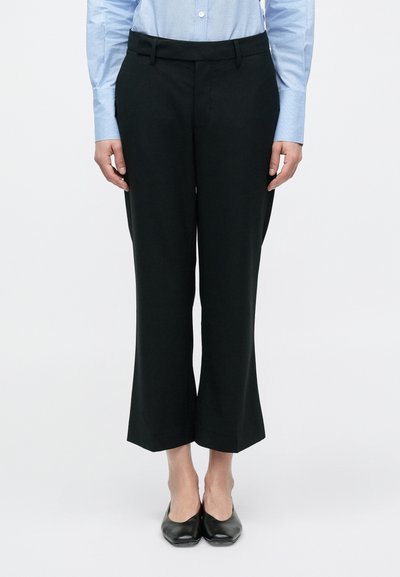 Pantalon évasé noir à texture lisse, avec une large ceinture et une coupe ajustée. Porté avec une chemise cintrée bleu clair.